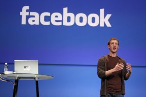 Facebook founder Mark Zuckerberg on stage with logo behind him on screen and a laptop on stage with him